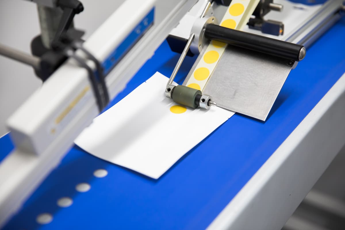 A yellow indicator dot being applied to a white label by a labelling machine applicator roller on a blue conveyor.