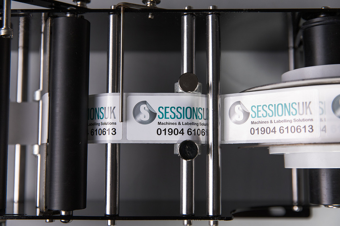 A web of Sessions UK labels passing between the black rollers of a labeller.